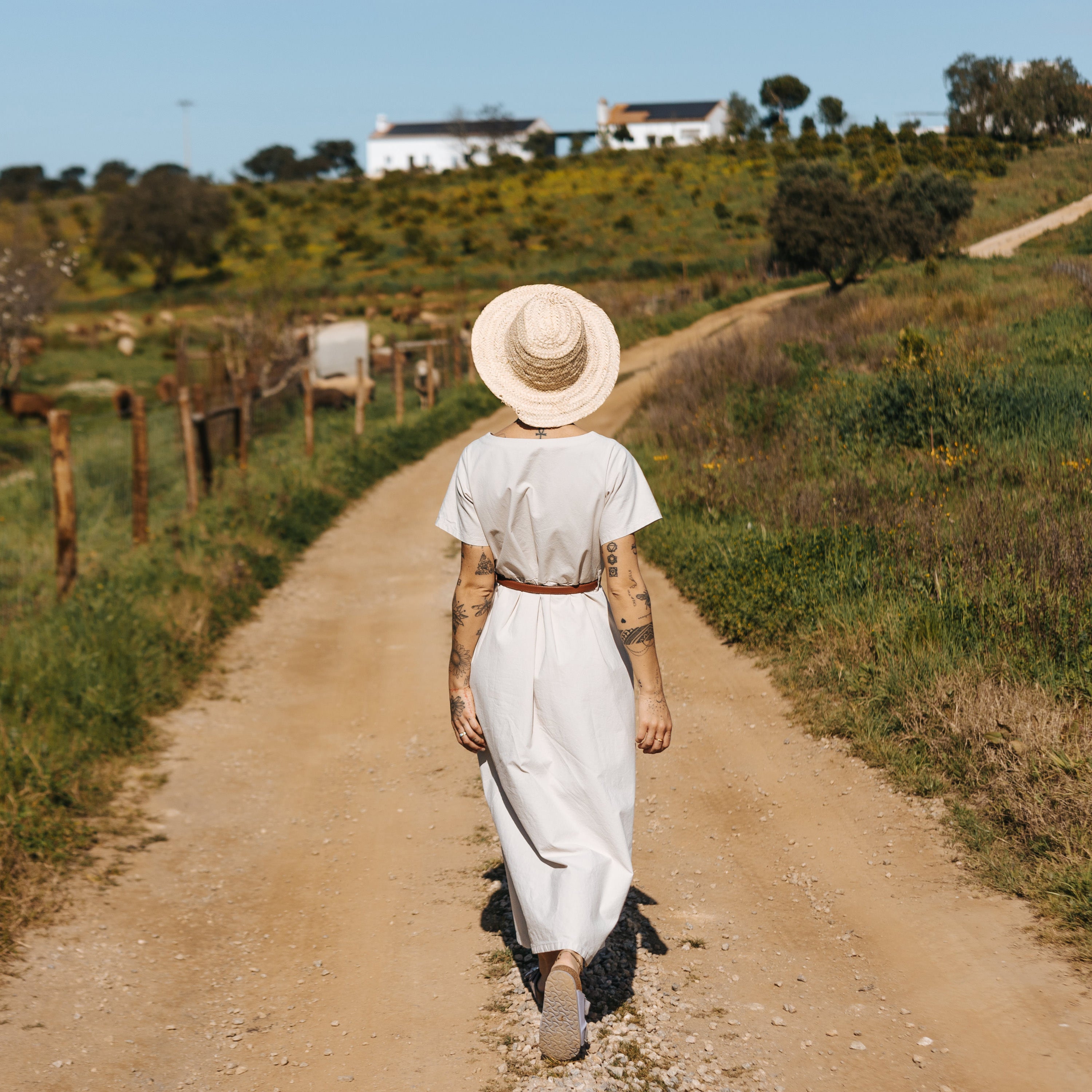 Image of a girl turned back, wearing a straw hat and a long white sustainable cotton dress, walking in a farm with meadows on both sides