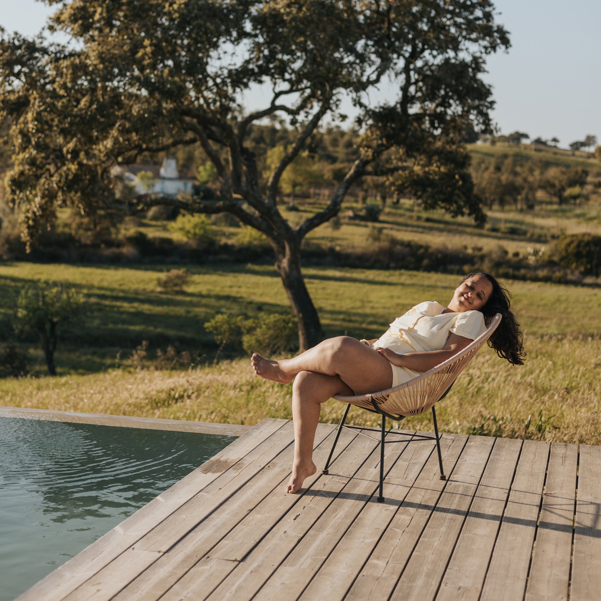 Mid-size tanned woman sitted in an outside chair taking a sun bath on the side of an ouside pool. She's wearing organic cotton made shorts and shirt.